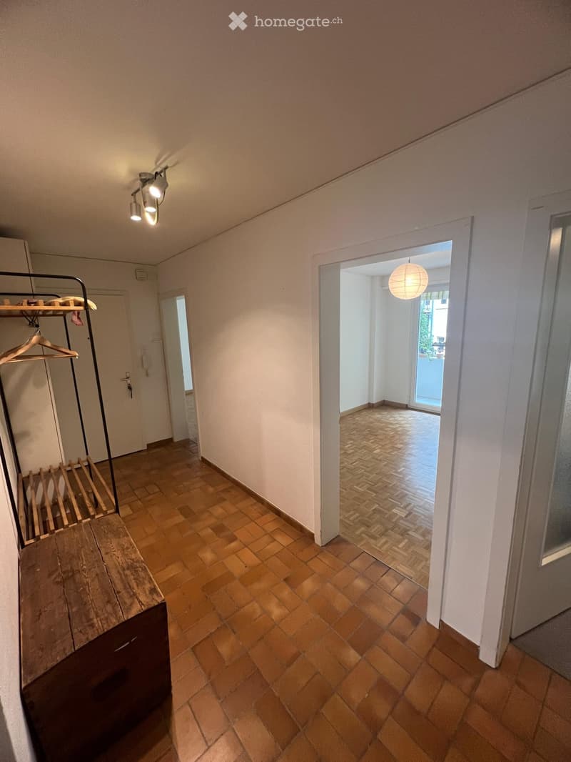 Empty hallway with tiled flooring, white walls, and recessed lighting. A coat rack with hangers is visible on the left, and an open doorway leads to another room with similar flooring.