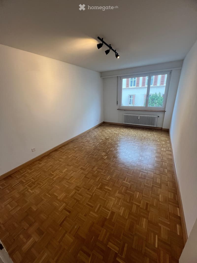 Empty room with wooden herringbone flooring, white walls, large window with blinds, radiator, and track lighting fixture.