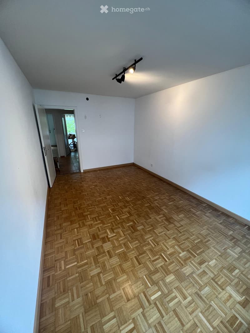 Empty room with light blue walls and herringbone wooden flooring. A single track light fixture is mounted on the ceiling, and an open doorway leads to another room with visible furniture.