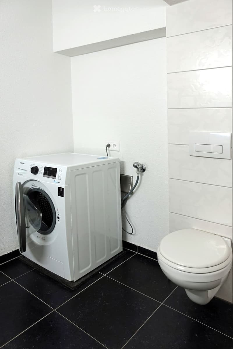 Modern bathroom with a white washing machine and toilet, black tiled floor, white walls, and a sleek design.