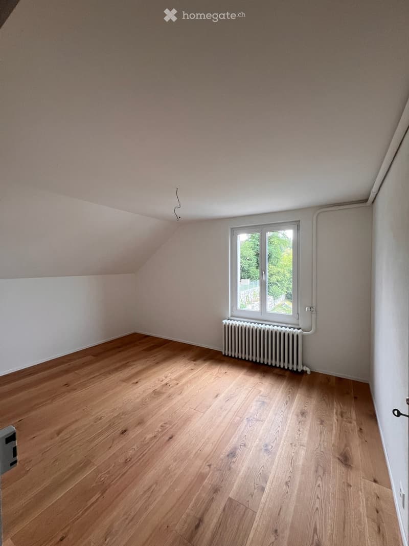 Empty room with wooden flooring, white walls, and a window with a radiator below it. The ceiling is slanted, typical of an attic space.