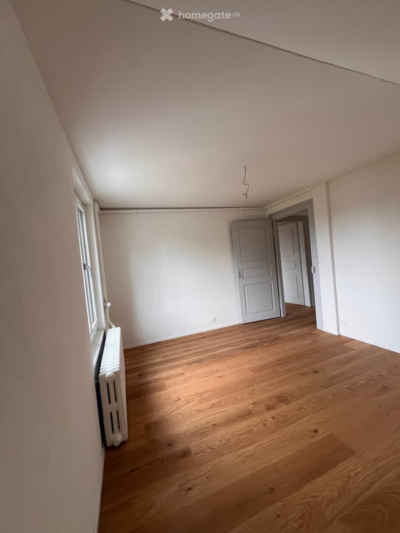 Empty room with wooden flooring, white walls, and a radiator on the left side. A door is visible at the back, leading to another room.