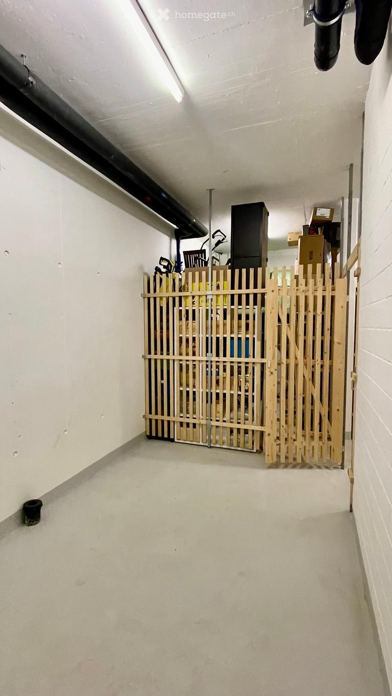Empty room with white walls and a concrete floor, featuring a wooden gate and some storage items behind it.
