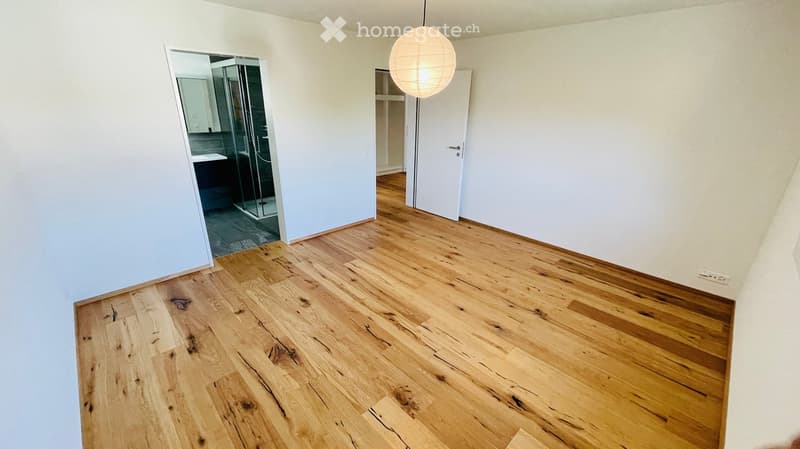 Empty room with light wooden flooring and white walls, featuring a hanging spherical light fixture and an open doorway leading to a bathroom area.