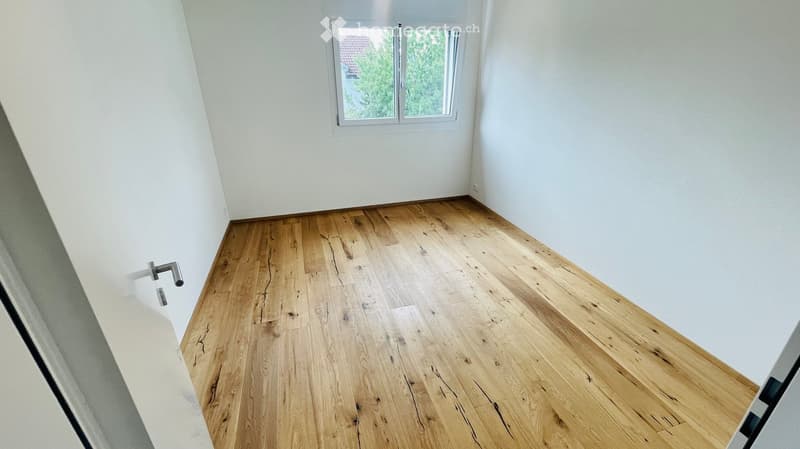 Empty room with light wooden flooring and white walls, featuring a single window with a view of greenery outside.