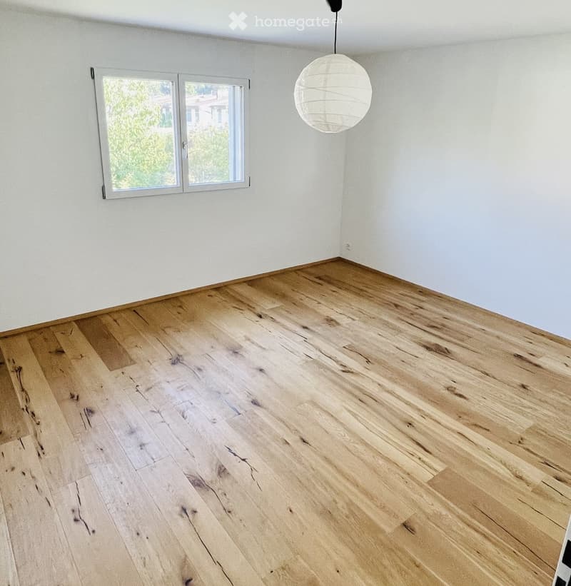 Empty room with light wooden flooring and white walls, featuring a large window and a hanging spherical light fixture.
