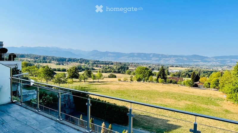 Balcony with a glass railing overlooking a scenic countryside with fields, trees, and distant mountains.