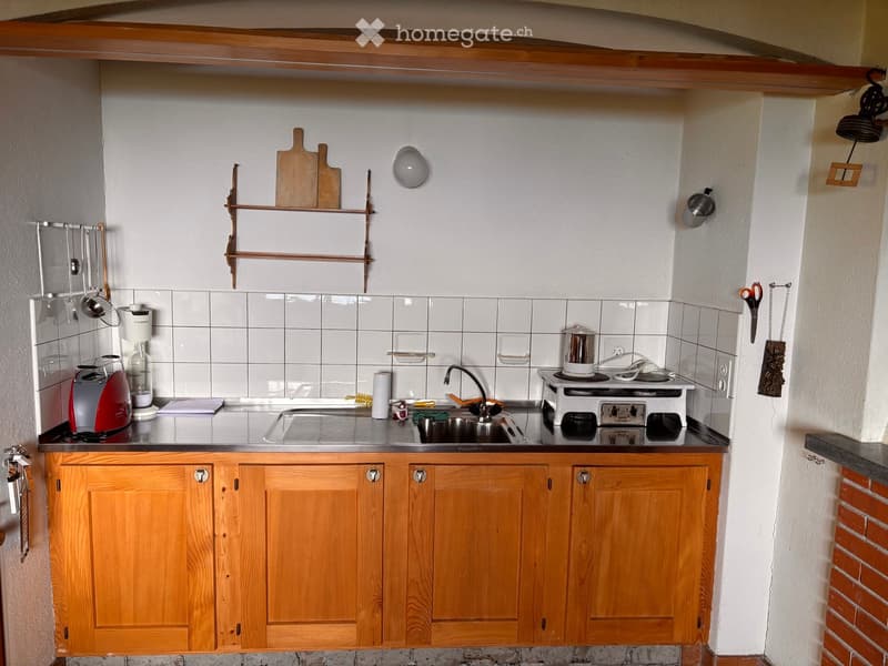 The image shows a small, well-organized kitchen with wooden cabinets, a stainless steel sink, a small stove, and various kitchen utensils and appliances neatly arranged on the counter and shelves.