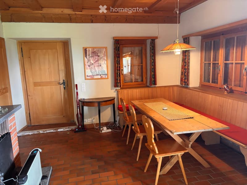 Cozy kitchen with wooden table, chairs, bench seating, and wooden cabinets. Features a hanging lamp, picture frame on wall, and a vacuum cleaner in the corner.