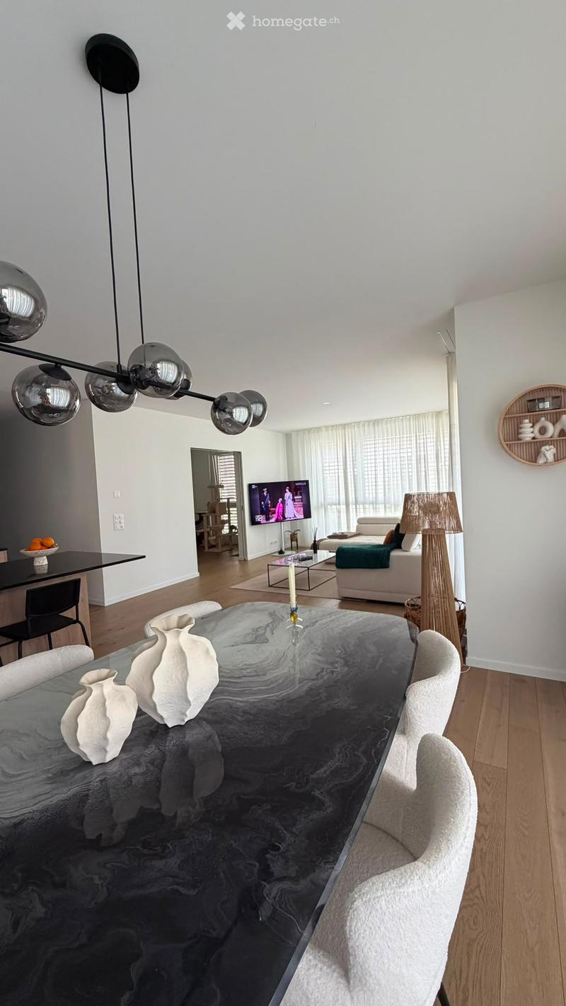 Modern living room with a large glass coffee table, white vases, a sofa, a television, and a floor lamp. The room features a sleek pendant light fixture and a dining area with a black table and chairs.