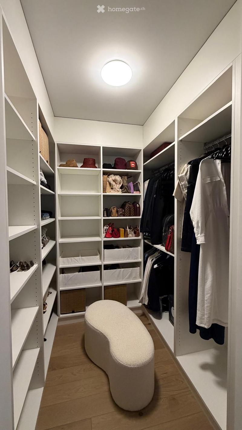 A modern, well-organized walk-in closet with built-in white shelving units, neatly arranged clothing, accessories, and a round, upholstered stool in the center.