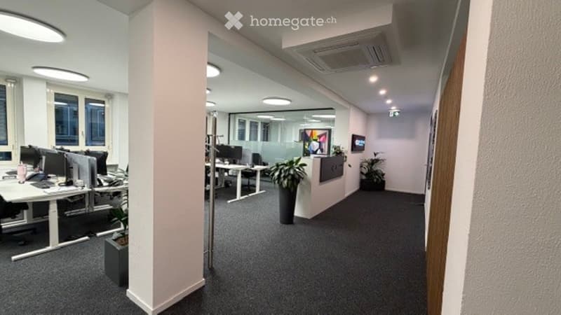 Modern office space with desks, chairs, computers, and plants. The area features a central column and large windows allowing natural light.