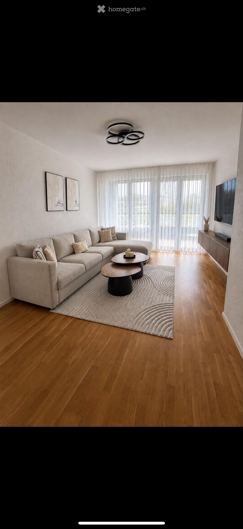Modern living room with a sectional sofa, coffee table, TV, and large windows with sheer curtains.