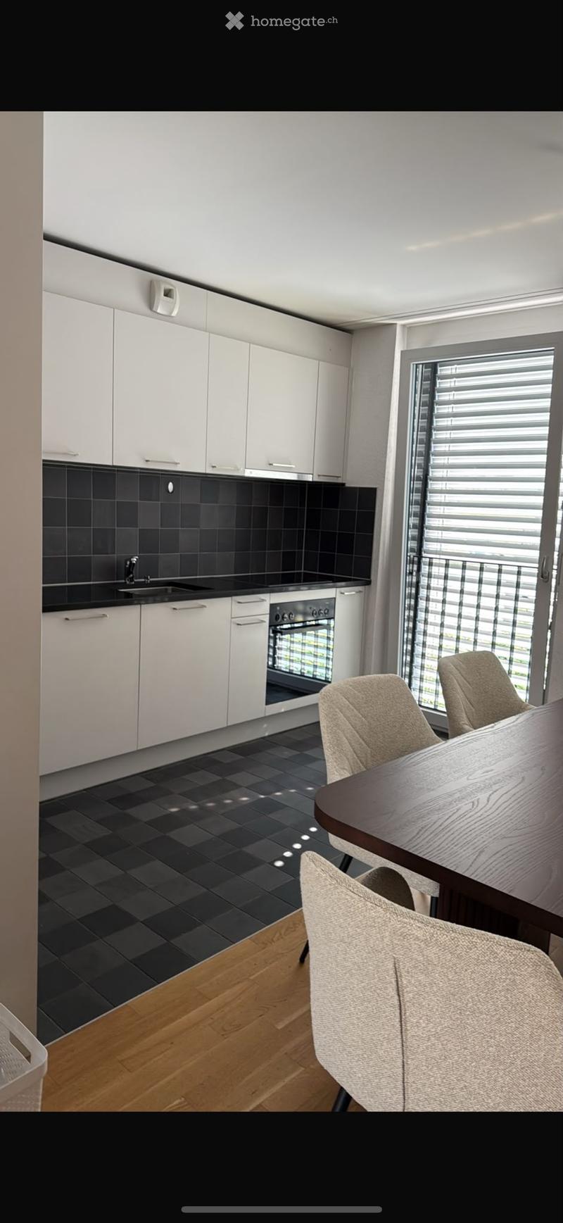 Modern kitchen with white cabinets, black tiled backsplash, and a dining area featuring a wooden table and beige chairs.
