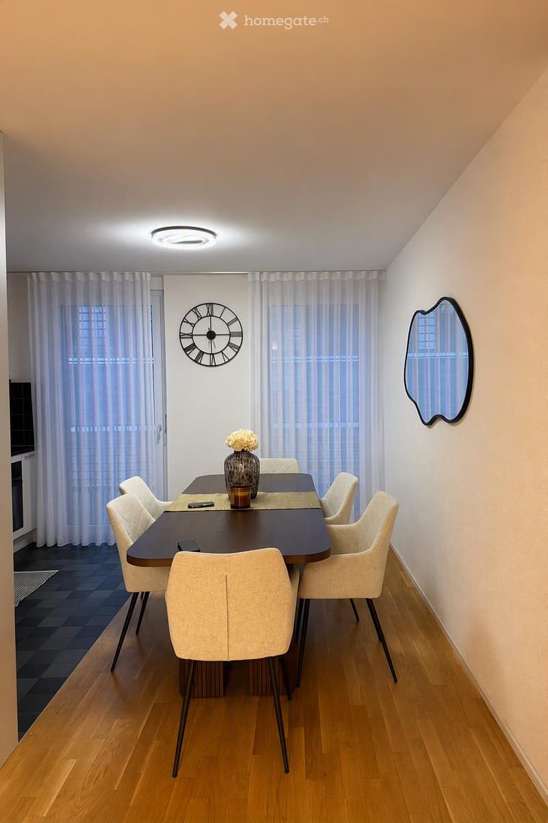 Dining room with a dark wooden dining table, six light-colored chairs, a vase with white flowers, a wall-mounted mirror, and a decorative clock. The room features wooden flooring and large windows with white curtains.