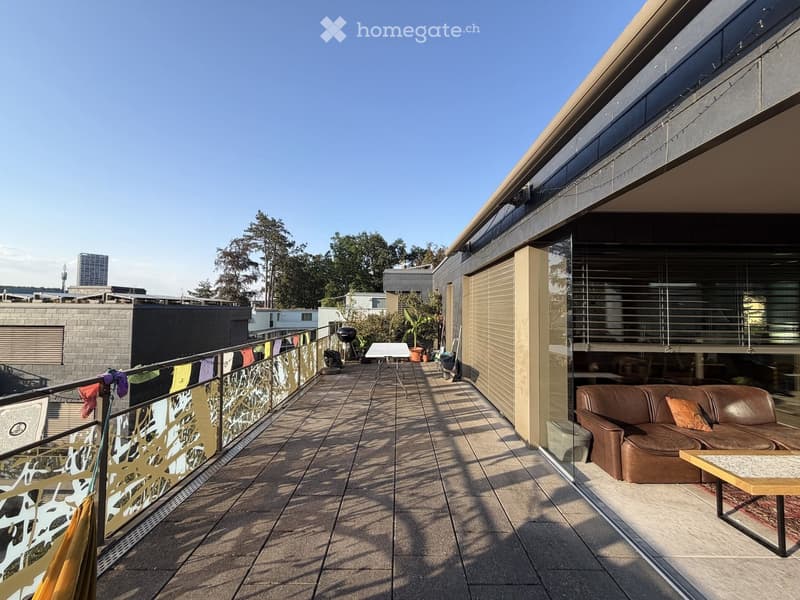 Balcony with modern glass railing, tiled floor, sofa, small table, barbecue, and potted plants. Offers a view of the city and surrounding greenery.