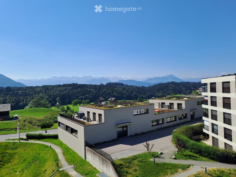 Modern multi-story building with flat roofs and large windows, surrounded by green fields and mountains in the background.