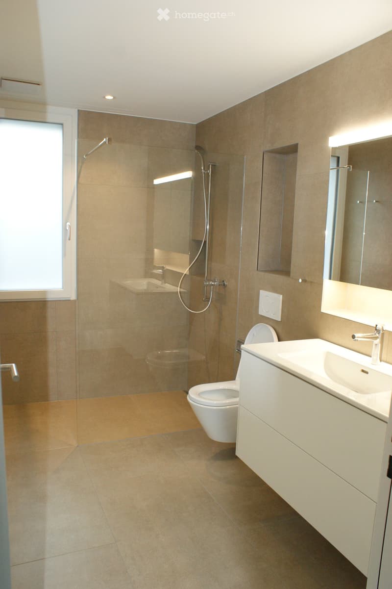 Modern bathroom with beige tiles, a white toilet, a sink with a mirror, and a glass-enclosed shower area.