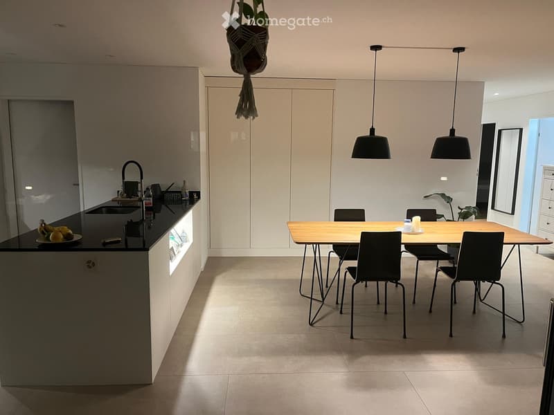 Modern kitchen with a black countertop, wooden dining table, and pendant lights. Includes a sink, faucet, and a hanging plant.
