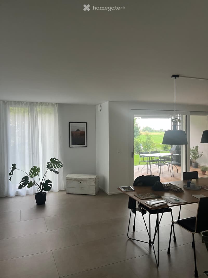 Bright dining area with tiled floor, large window with white curtains, and a view to a patio with outdoor furniture.