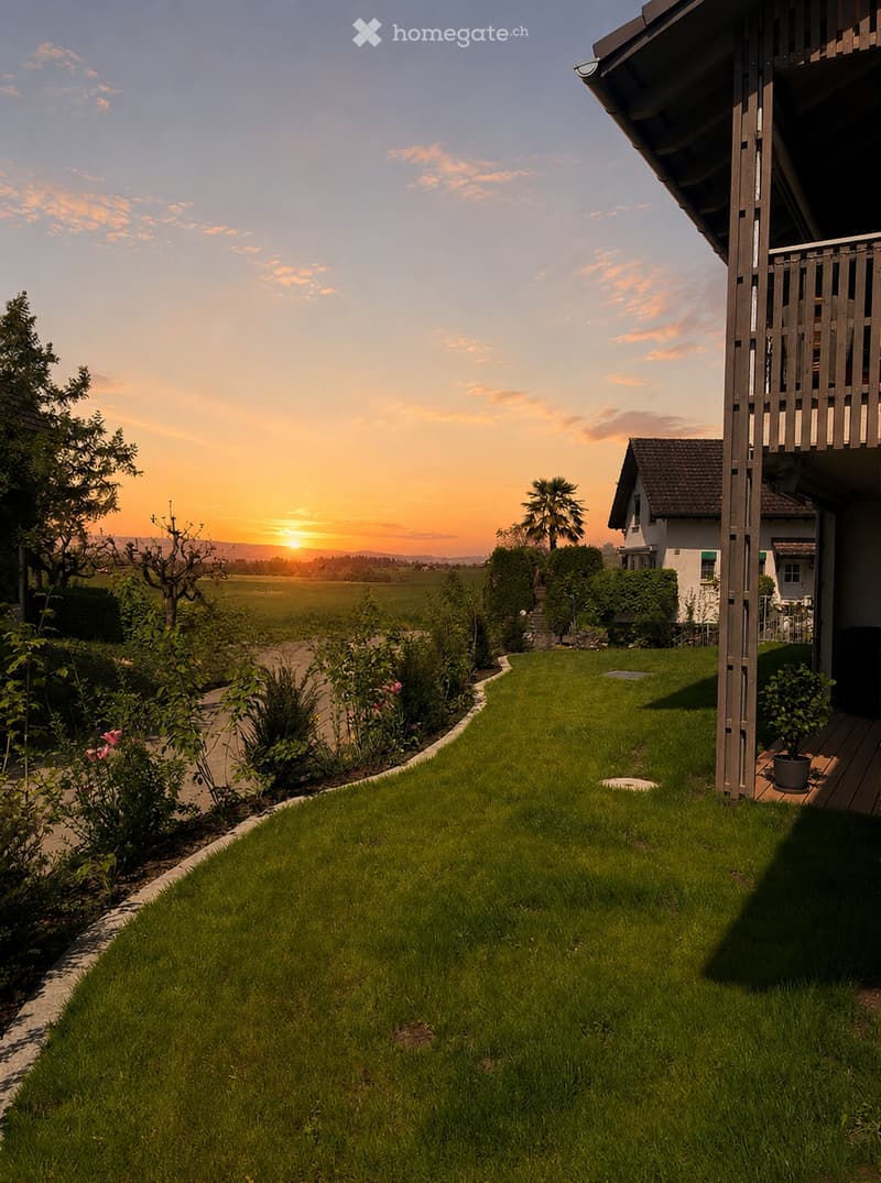 Well-maintained lawn with a curved pathway, surrounded by shrubs and flowers, leading to a house with a balcony and a view of a sunset over a field.