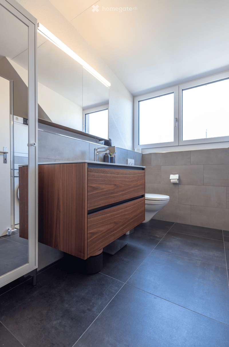 Modern bathroom with dark tiled floor, wooden vanity cabinet, sink, faucet, and toilet. Large windows provide natural light.