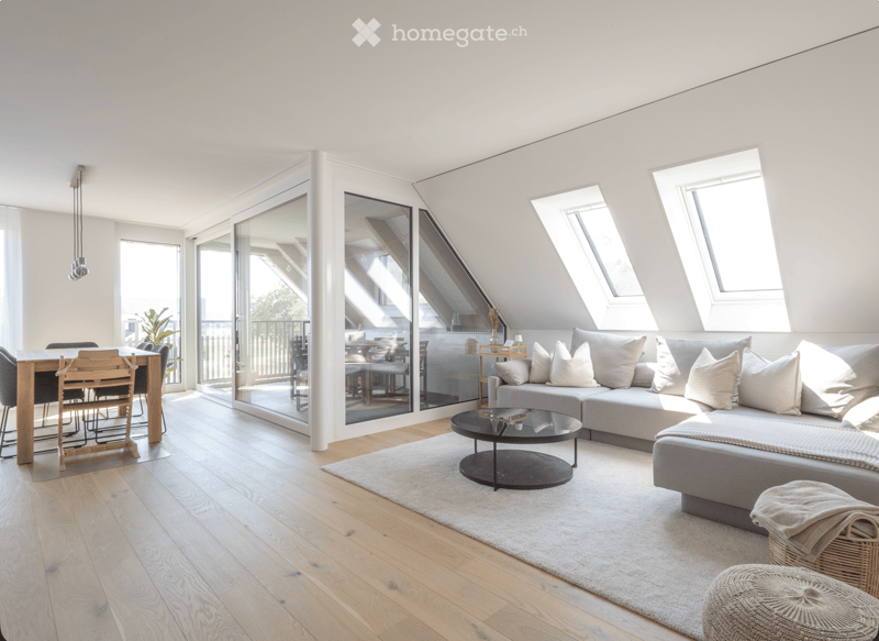 Modern living room with a grey sectional sofa, coffee table, ottoman, and a dining area with a wooden table and chairs. Large windows and skylights provide ample natural light.