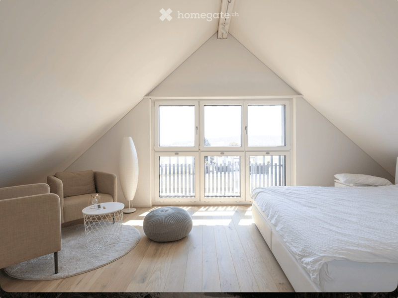 Bright, modern bedroom with wooden flooring, large windows, a bed, sofa, chair, round table, and floor lamp.