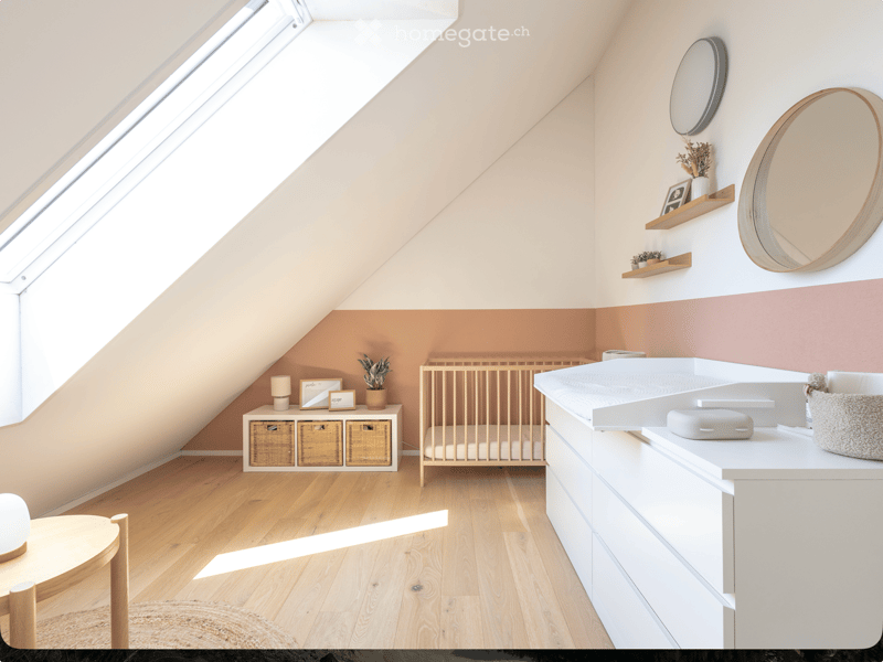 Cozy attic bedroom with wooden flooring, slanted ceilings, and large skylight. Features a crib, changing table, round mirror, shelves with decor, and a small round table.