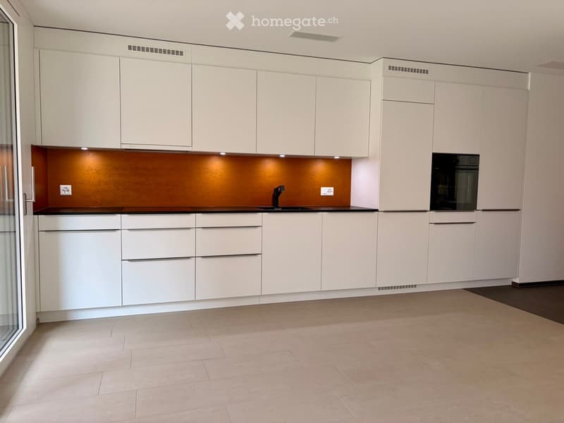 Modern kitchen with white cabinets, a dark countertop, and built-in appliances including a sink and oven. The floor is tiled, and the space is well-lit with integrated lighting.
