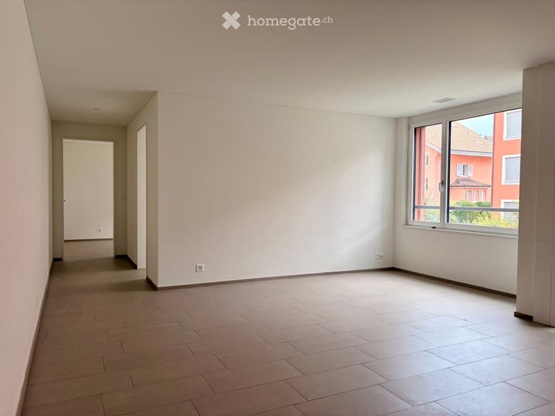 Empty room with light-colored walls, tiled floor, and large window with view of neighboring building.