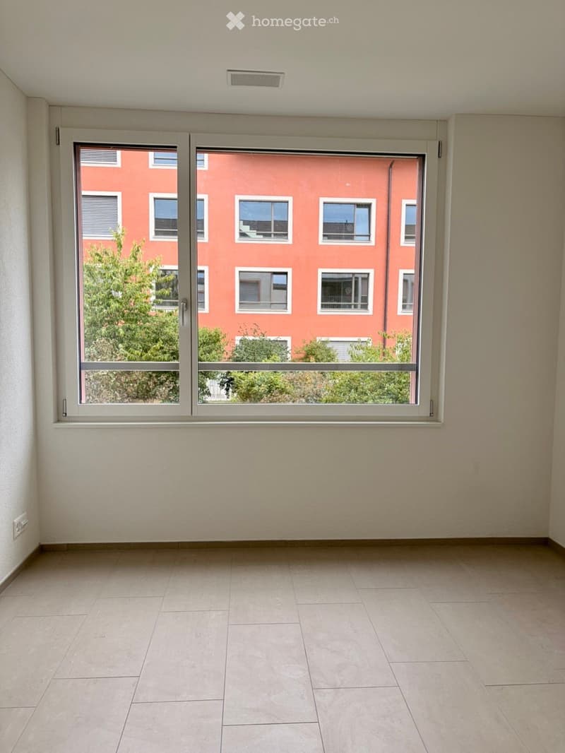 Empty room with tiled floor, large window with view of a red building and greenery outside.