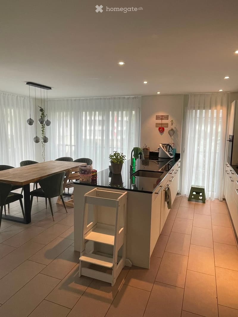 Modern kitchen with a wooden dining table, black chairs, a kitchen island, and pendant lights. The island features a sink, faucet, and a small plant. A green stool is placed near the island.