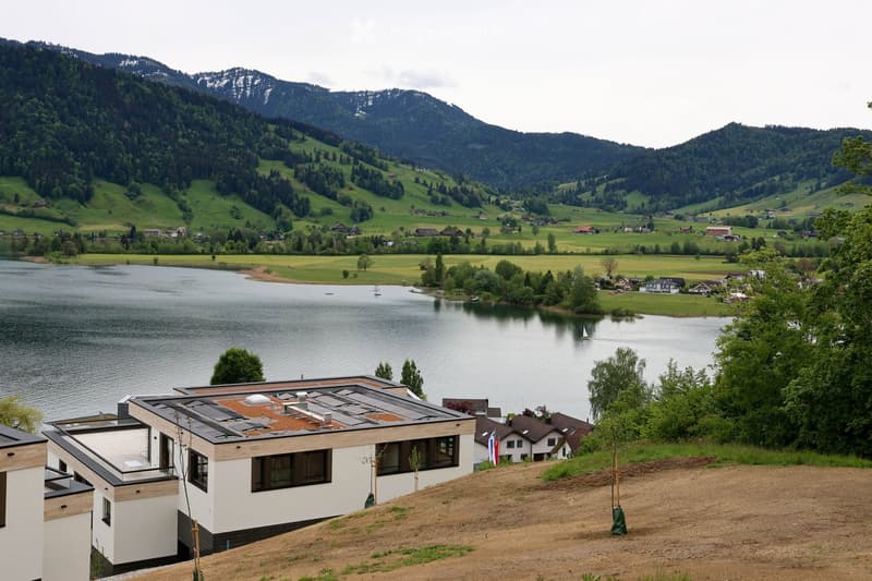 Modern house with a flat roof and solar panels, situated on a hillside overlooking a lake and surrounding mountains.