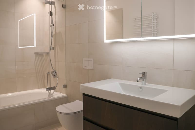 Modern bathroom with a bathtub, toilet, sink, and shower. Features a large mirror with built-in lighting and sleek, contemporary fixtures.