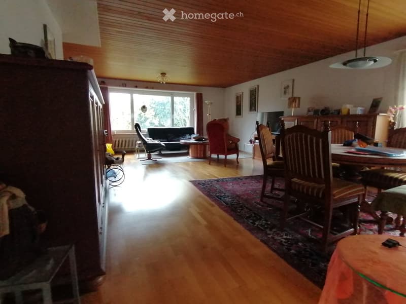 A spacious, well-lit living and dining area with wooden flooring, a large window, a sofa, chairs, a dining table, and a cabinet.