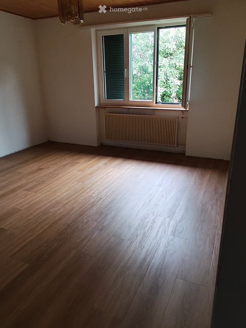 Empty room with wooden flooring, large window with shutters, radiator below the window, and visible ceiling beams.