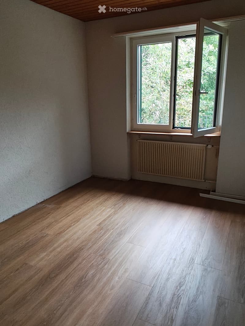 Empty room with wooden flooring, white walls, and a large window with a radiator below it.