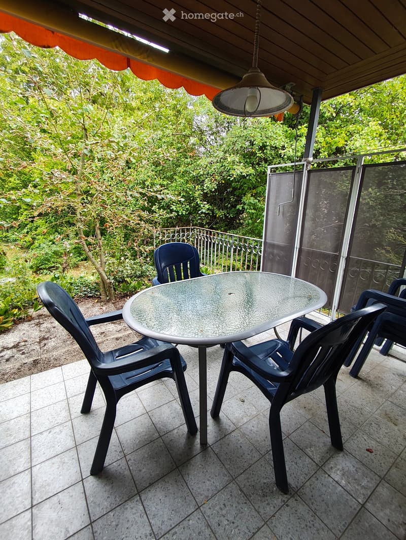 Balcony with a round glass table and four black plastic chairs, surrounded by lush greenery and a metal railing.