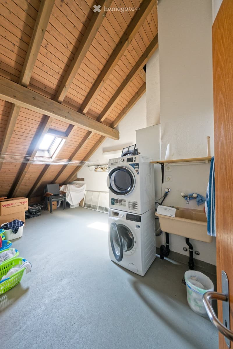 The room features a washing machine, a chair, a sink with a faucet, and a basket, with exposed wooden beams on the ceiling and a skylight letting in natural light.