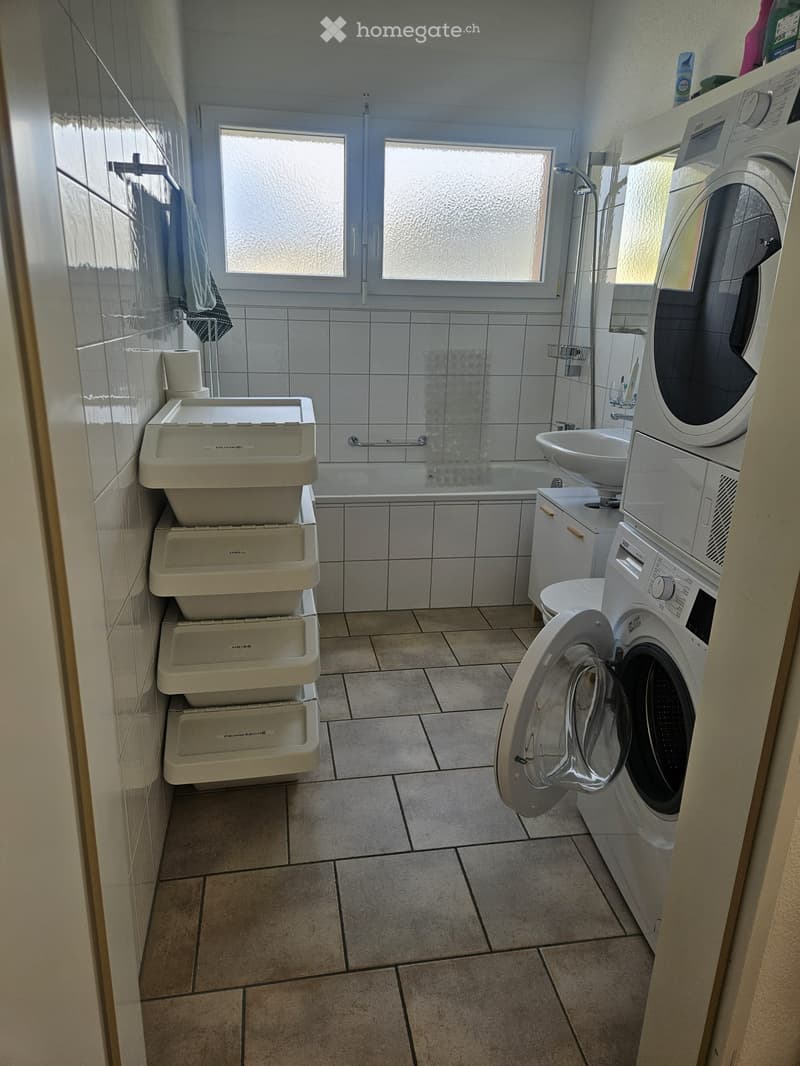 Bathroom with tiled walls and floor, bathtub, sink, toilet, washing machine, and a towel hanging on the wall.