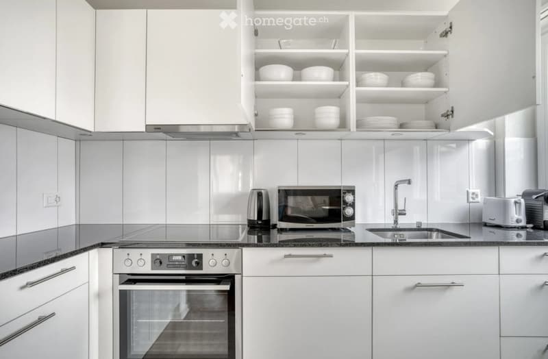 Modern kitchen with white cabinets, stainless steel appliances, and a black countertop. Visible items include an oven, microwave, sink, faucet, toaster, and dishes in the cabinets.