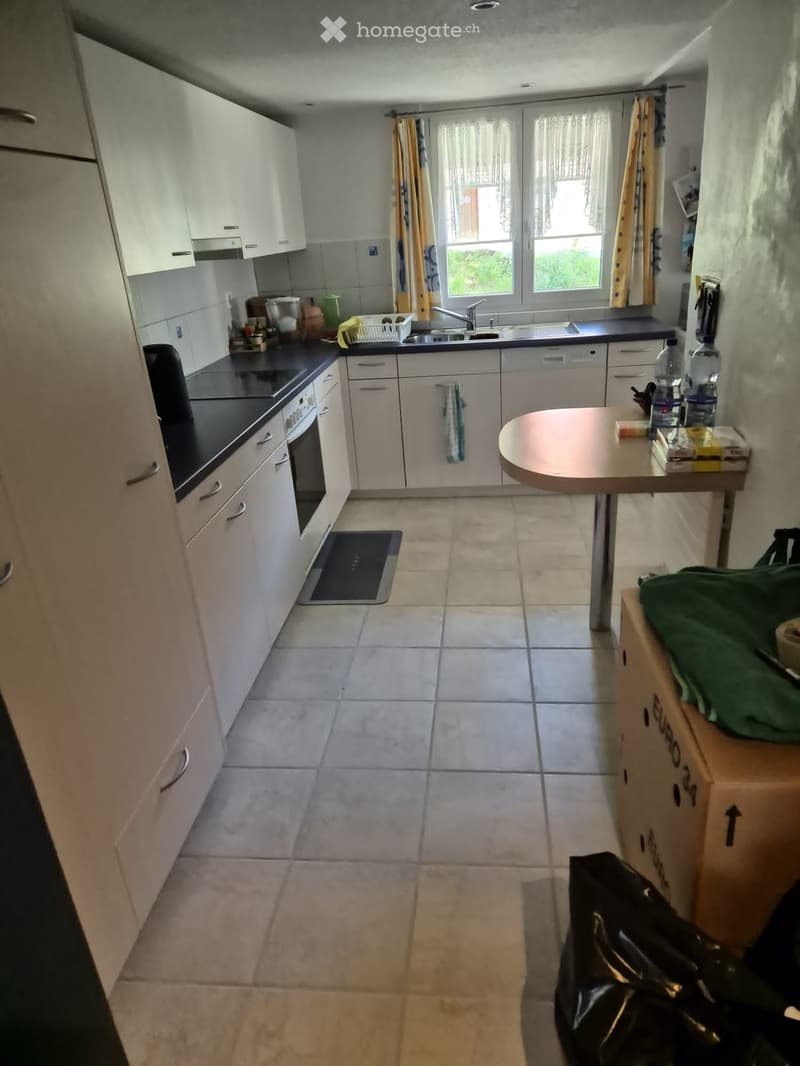 Modern kitchen with white cabinets, black countertop, sink, and a small table with items on it. The floor is tiled, and there are boxes and a bag in the corner.