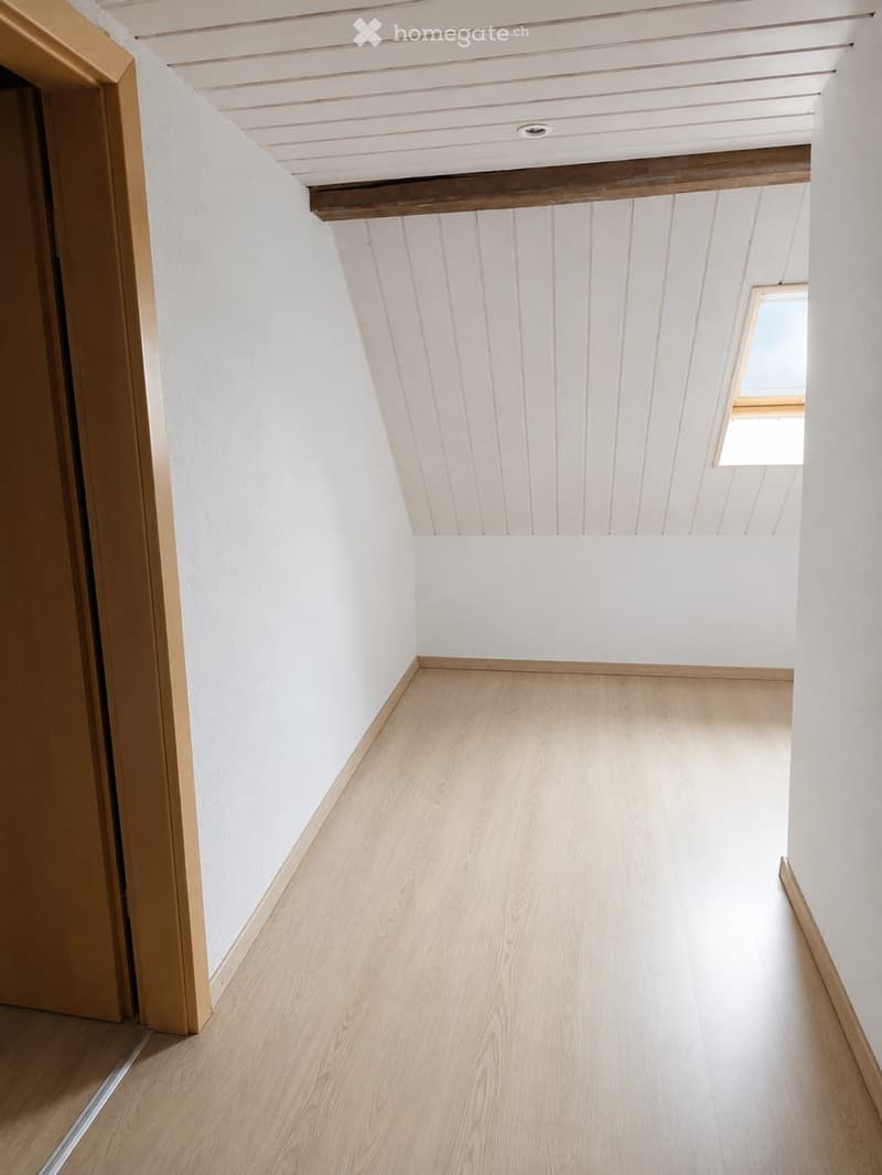 Empty hallway with light wooden flooring, white walls, and a slanted white ceiling with exposed wooden beams. A small window at the far end allows natural light to enter.