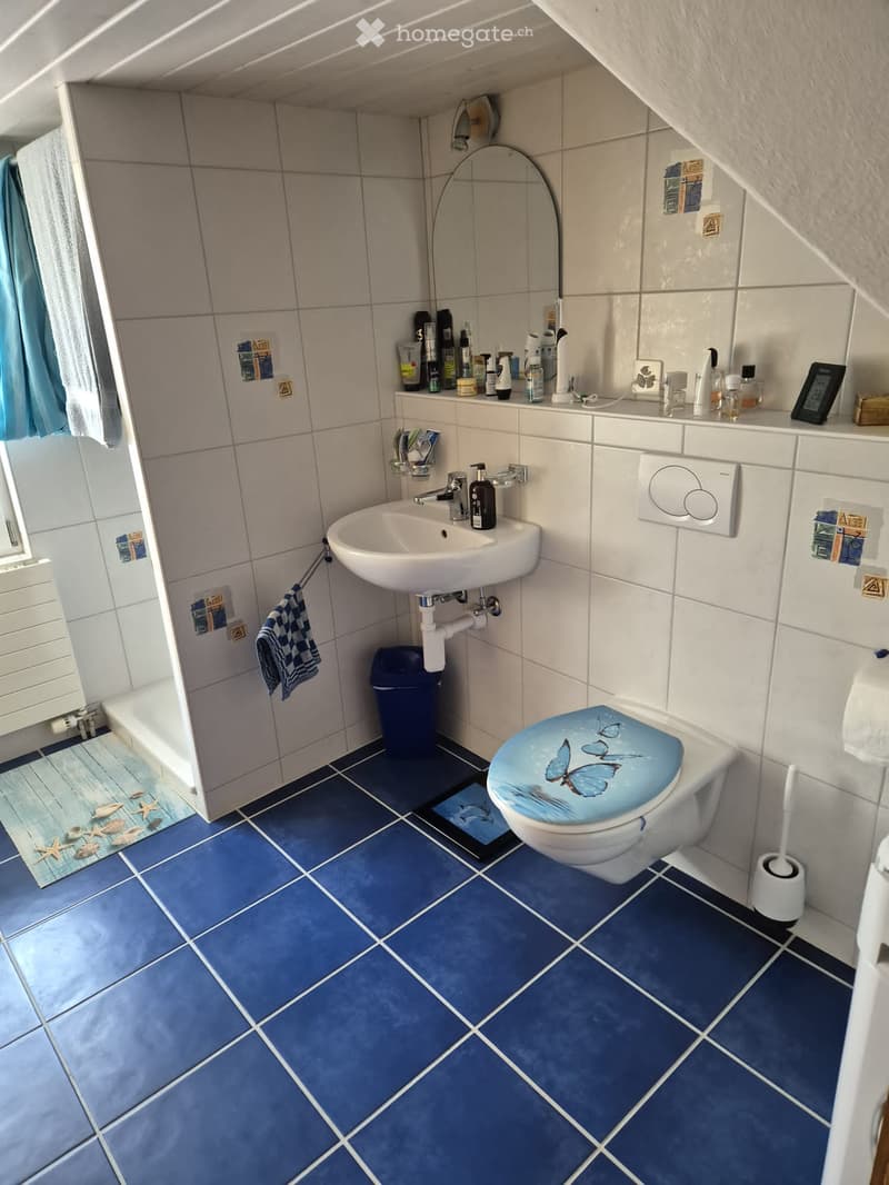A small bathroom with blue tiled floor and white tiled walls, featuring a sink, toilet with a butterfly seat cover, mirror, towel, and a small cabinet with toiletries.