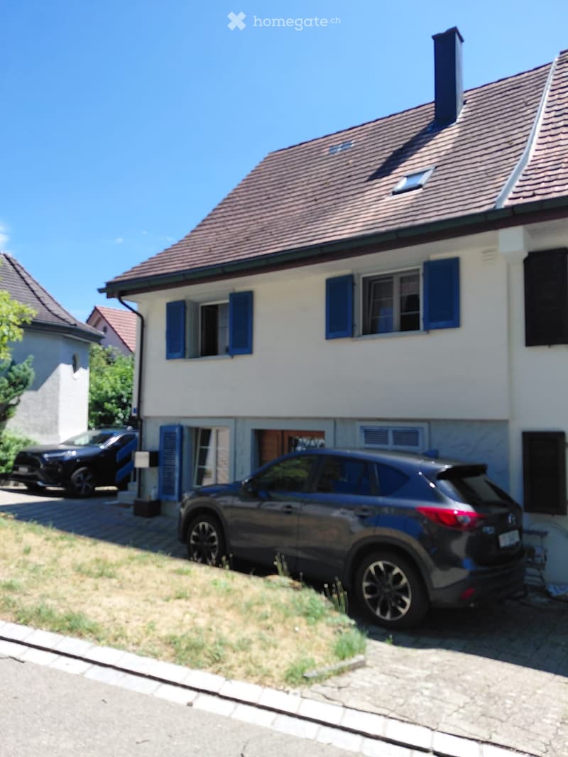 Two-story house with white walls, blue shutters, and a red-tiled roof. Two cars are parked in the driveway.
