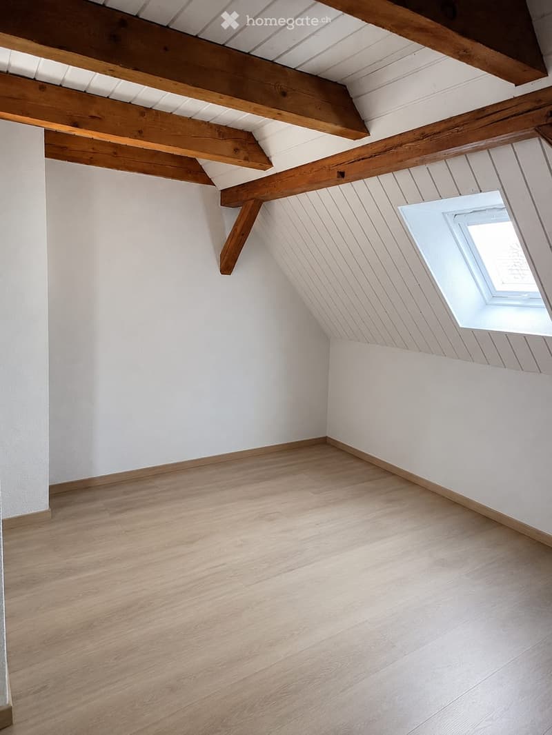 Empty attic room with wooden floor, white walls, exposed wooden beams, and a skylight window.