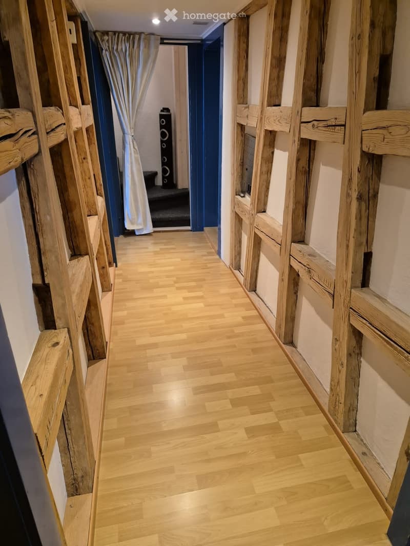 Wooden-floored hallway with exposed wooden beams and built-in wooden shelves on both sides. A curtained doorway leads to another room with a visible speaker system.