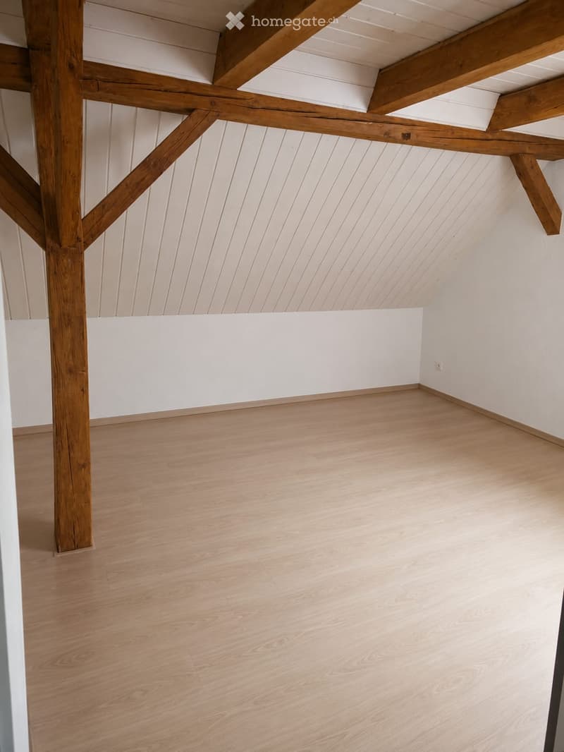 Empty room with light wooden flooring, white walls, and exposed wooden beams on the ceiling.