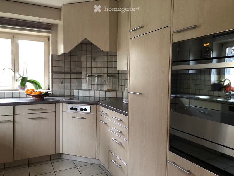 Modern kitchen with light wood cabinets, stainless steel appliances, and a black countertop. Features an oven, sink, and various kitchen items on the counter.